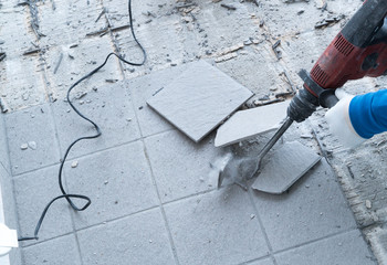 construction worker using a handheld demolition hammer and wall breaker to chip away and remove old floor tiles during renovation work - obrazy, fototapety, plakaty