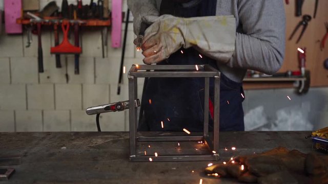 Slow motion shot of unrecognizable metalworker in protective gloves using welding torch to join sides of metal cube standing at workbench, sparks flying around