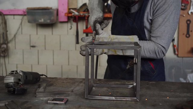 Closeup shot of unrecognizable metalworker in protective mask and gloves using welding torch to join sides of metal cube standing at workbench, sparks flying around in slow motion