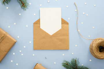 Empty blank card in kraft paper envelope on pastel blue background decorated with confetti star, gift box, wrapping paper and fir tree branches. Christmas, New Year, winter holiday invitation mockup.
