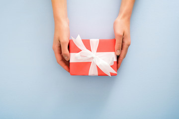 Female's hands holding red gift box with white ribbon bow on pastel blue background. Christmas, New Year, Valentine's day and birthday concept. Minimal flat lay style composition, top view, overhead