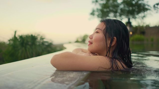 Outdoors holidays lifestyle shot on young beautiful and relaxed Asian Chinese woman enjoying at tropical resort swimming pool with a view to tropical landscape and palm trees on Summer sunset