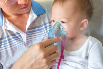 Asian father helping his toddler son with inhalation therapy by the mask of inhaler. Sick little kid with respiratory problem with oxygen mask breathes through nebulizer at doctor clinic / hospital