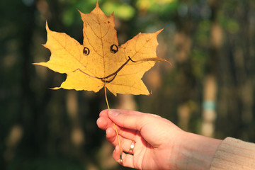 Golden autumn time. Young woman is holding a single maple leaf with cheerful expression facial (painted smile) Fallen leaves in old city park, background