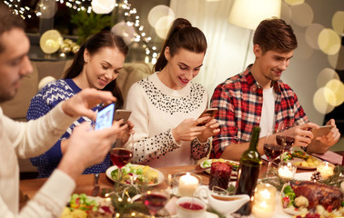 holidays and celebration concept - happy friends with smartphones having christmas dinner at home