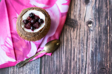 Berry dessert with yogurt in a cup of coconut on a wooden background. Blackberries, raspberries, blueberries, black currants. Pink napkin