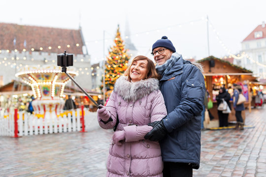 technology, winter holidays and people concept - happy senior couple taking picture by smartphone on selfie stick at christmas market on town hall square in tallinn, estonia