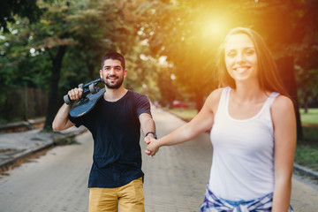 young happy couple holding arms walking in park holding skateboard
