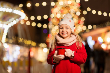holidays, childhood and people concept - happy little girl with cup of tea at christmas market in winter evening