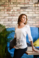 young brunette in a white T-shirt is standing near a table in a cafe with a phone in her hands, she is waiting for her friend or colleague to meet. Not a forced environment. Modern interior.