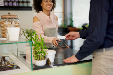 Smiling mixed race female pastry shop employee billing bill while Caucasian man holding credit card.