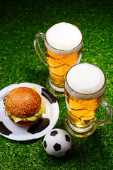 Two glasses of beer, hamburger and soccer ball on green grass.