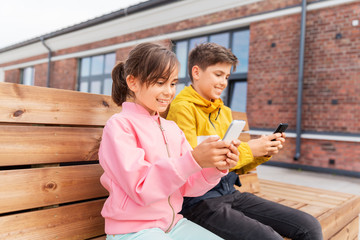 childhood, technology and people concept - happy children or brother and sister with smartphones sitting on wooden street bench outdoors