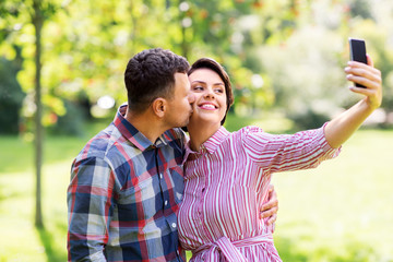 people, technology and relationships concept - happy couple in summer park taking selfie by smartphone