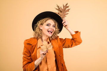 Stylish woman with autumn leaves in orange jacket. Beautiful model girl with autumnal leaves. Woman autumn fashion. October. Fashionable woman with autumn leaves in hand. Autumn girl in black hat.