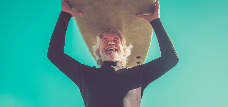 Happy Senior With Surftable On His Head Is Smiling And Laughing - Old And Mature Man Having Fun Surfing With A Black Wetsuits - Active Retired Adult Doing Activity Alone