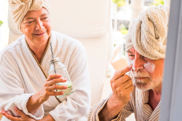 couple of seniors sitting together at hotel with spa and beauty tratament - close up of man putting some cream on his face with her wife looking at he