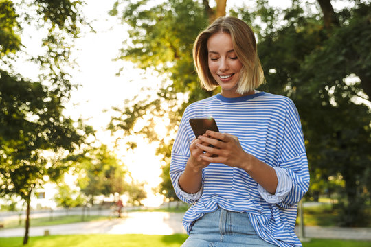 Image of smiling woman typing on cellphone while resting in park