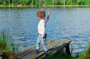 Fototapeta na wymiar little girl child with curls stands on the shore of a forest lake on a wooden pier and throws stones into the water in summer day.