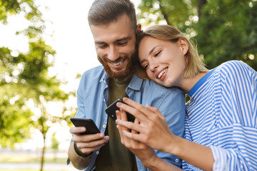 Image of smiling couple using cellphones while resting in green park