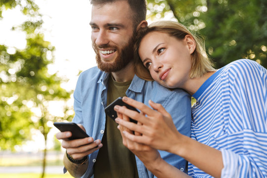 Image of young romantic couple using cellphones while resting in park