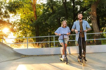 Image of caucasian happy couple smiling while riding e-scooters
