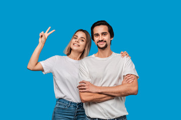 Happy couple of a young blond woman and brunet bearded man with mustaches in white t-shirts and blue jeans posing isolated over blue background. Concept of an ideal couple.