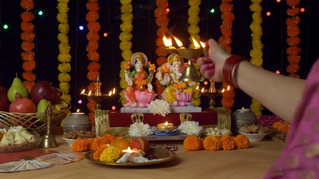 Over the shoulder shot of a female worshipping Lord Ganesha and Goddess Laxmi - Diwali puja. Indian female praying in front of Ganesh Ji and Lakshmi Ji with a burning oil lamp - bokeh floral lights...