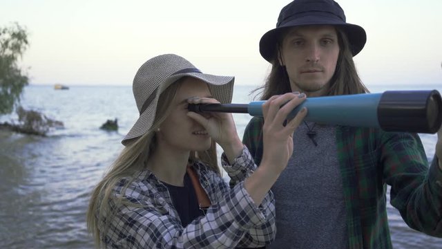 Young caucasian tourist couple in shirts and hats looking throw telescope and chatting on the background of sea or lake.