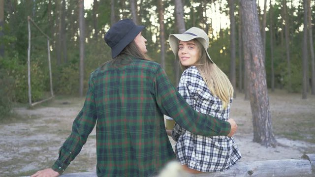Back view of tourist couple in plaid hipster shirts and hats sitting in the forest. Young caucasian girl is drinking tea or coffee from the yellow cup resting next to her boyfriend