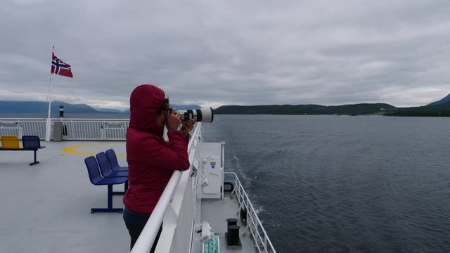 Tourist woman on windy cruise ship deck enjoying fjord view, taking photo with camera. Ferry ride route Bognes - Lodingen to Lofoten islands Norway. Tourism vacation and travel