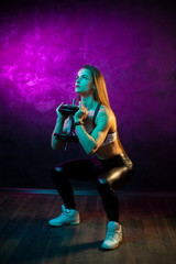 Focused young woman fitness model doing squats with professional dumbbells in neon lights silhouette in the studio.