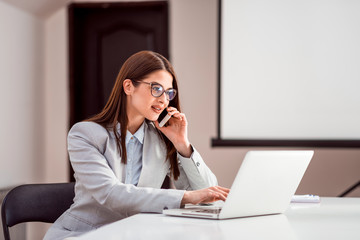 Cute young female adult with glasses talking on phone while working on laptop.
