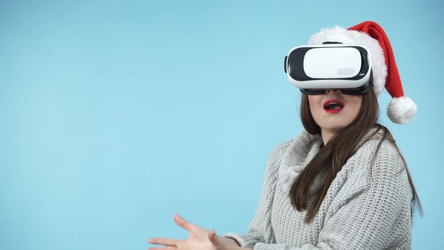 Woman wearing santa claus hat exploring space with virtual reality goggles headset. Amazed girl watching 3d film tour in vr glasses box. New generation cyber christmas concept, on blue