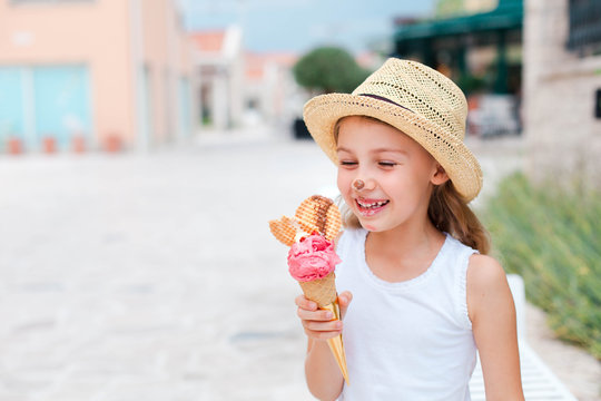 Fototapeta Funny kid is eating ice cream. Little girl is enjoying pink italian gelato. Happy cute child in straw hat is smiling and tasting delicious food in town streets. Lifestyle moments, summer traveling.