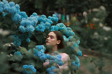 Tender fashion brunette girl with bright makeup wearing pink t-shirt standing among blue flowers with closed eyes
