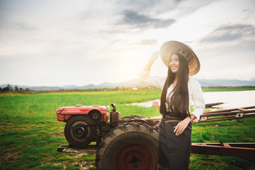 Asian Beautiful woman in Thai local dress standing nearby mini tractor with field and lake...