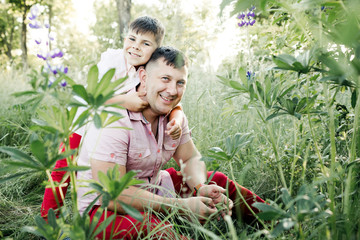 a father sits among greenery lawn, and a son hugs his behind