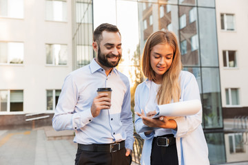 Cheery man and woman businesspeople outside at the street near business center holding clipboard with documents plans and cup of coffee.