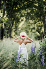 a woman stands turned a back and holds for her hat among greenery lawn