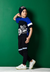 Teenager in sportswear headwear, sweatpants and t-shirt and sneakers is standing at the wall and looks back cool posing