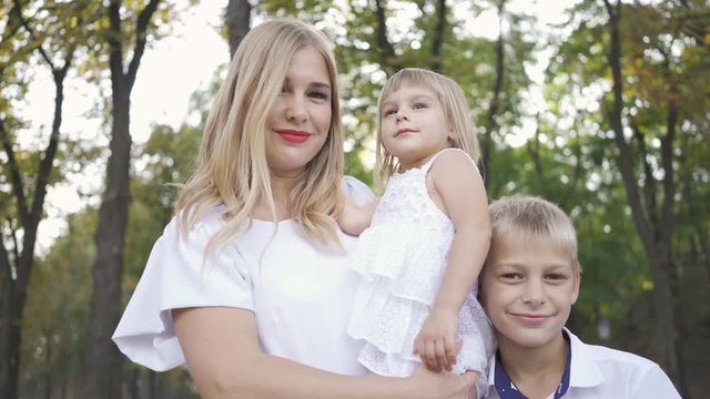 Attractive blond woman holding baby daughter in her arms while older son hugging mother smiling. Portrait of happy family with two kids looking at the camera