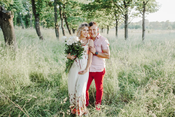 a man hugs a beautiful pregnant woman with peonies among greenery lawn