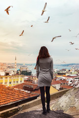 A girl stands on the roof of a house in Istanbul with her full back against the backdrop of a beautiful panorama of the city with flying gulls.Young woman admires the panorama of Istanbul
