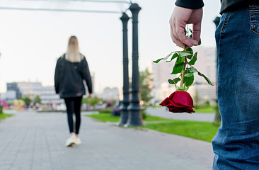 View of the red rose flower in the hand of the guy on the background of the girl leaving him in the city Park, square. Separation, resentment, quarrel, dislike, unloved