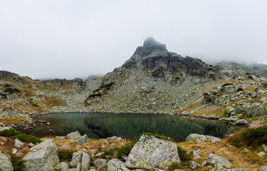Panoramic view of high mountain lake surrounded by rocks and stone blocks, behind it a rocky peak covered with mists, The Quiet lake ( Tihoto ezero ),  Rila National Park, Bulgaria.