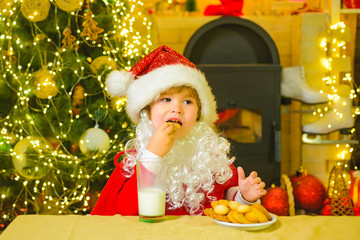 Fototapeta na wymiar Santa boy child eating cookies and drinking milk. Santa Claus - bearded funny child. Santa in home. Santa Claus takes a cookie on Christmas Eve as a thank you gift for leaving presents.