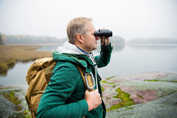 Mature man exploring Finland in the fall, looking into fog through binoculars. Hiker with big backpack standing on mossy rock. Scandinavian landscape with misty sea and autumn forest.