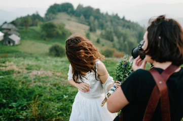 Beautiful bride in a white dress on a green field in the mountains, on the grass. Wind driveth brown hair. A photographer takes pictures of the bride. Wedding ceremony. Hipster. Rustic. Happy.