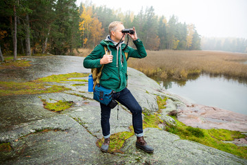 Mature man exploring Finland in the fall, looking into fog through binoculars. Hiker with big backpack standing on mossy rock. Scandinavian landscape with misty sea and autumn forest.
